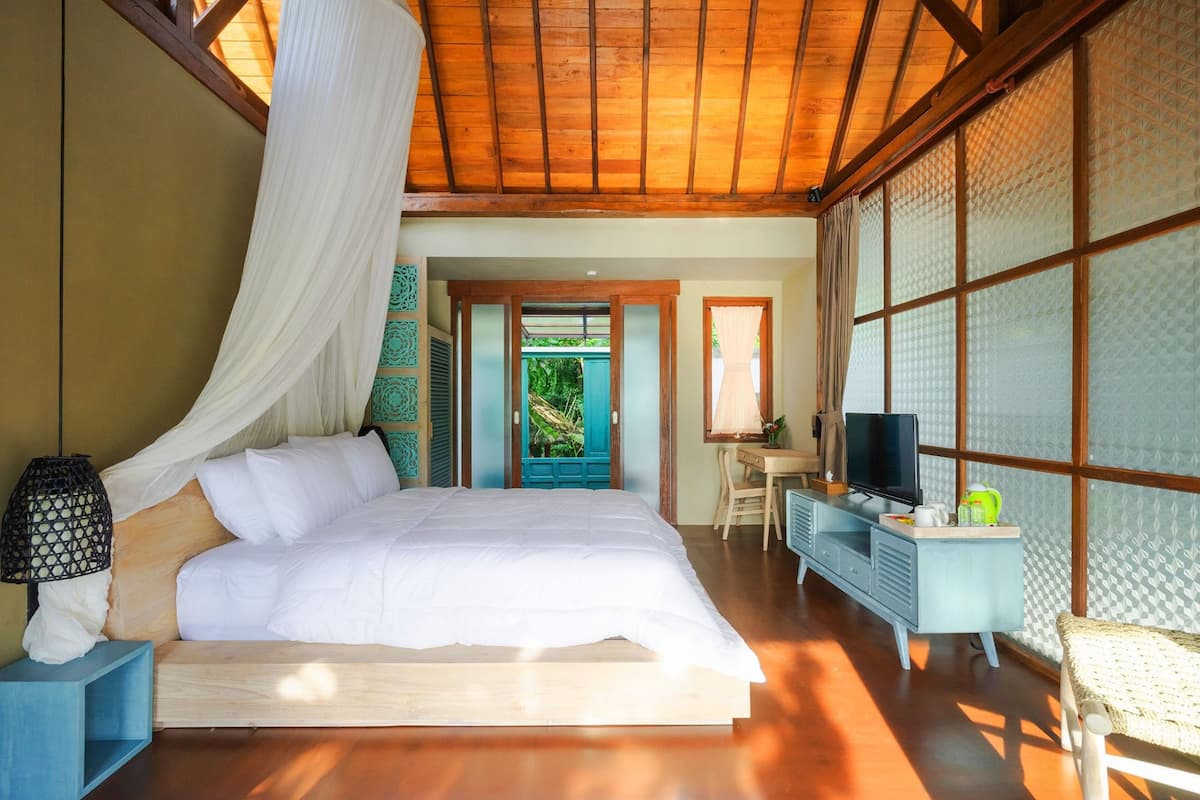 A bedroom with a large bed, a desk, a television, and a view of greenery outside.