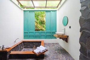 A bathroom with a stone floor, a wooden bench with towels, a sink with a bowl, and a window with green shutters. - Nayaka Living Ubud (Ubud)