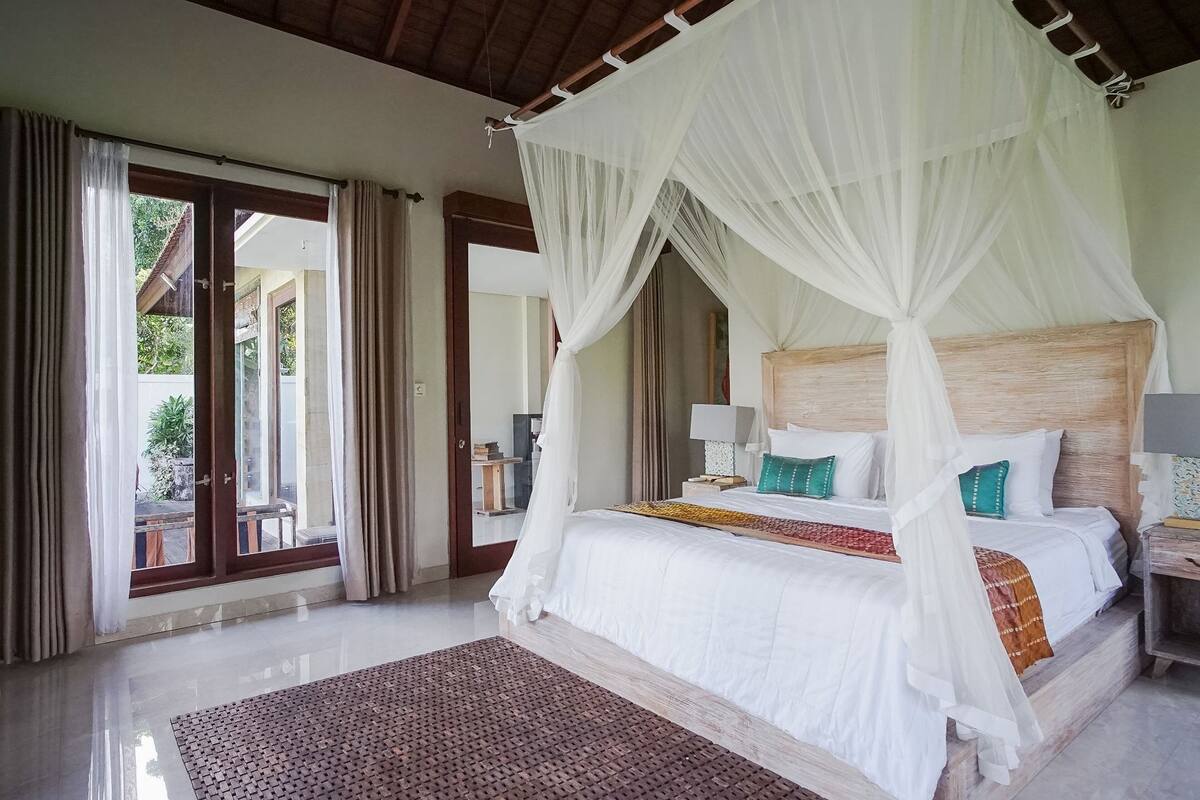 A spacious bedroom with a large bed, white curtains, and a view through the glass doors.