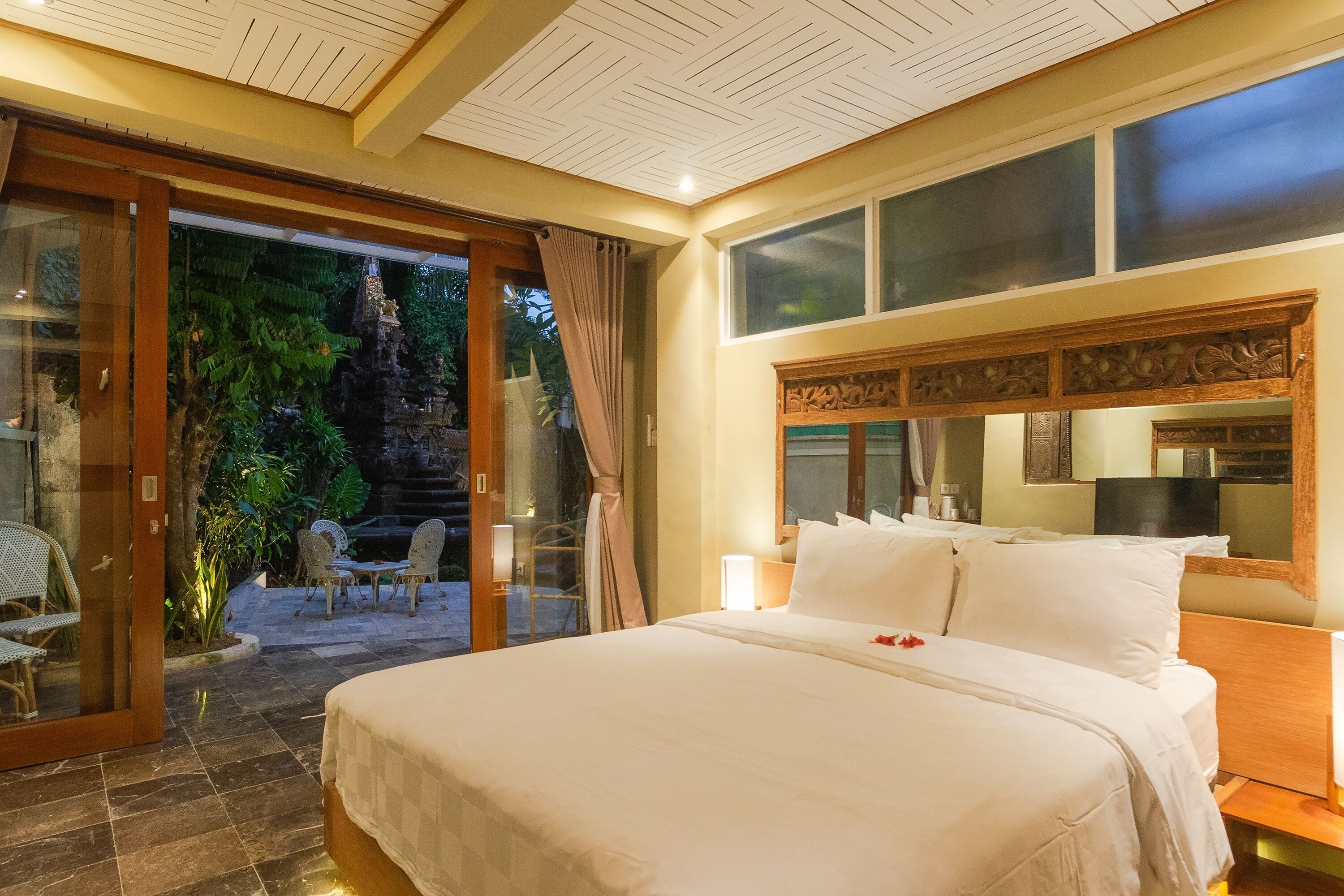Habitación de hotel con una cama grande, cabecera de madera, puerta de vidrio que da a un espacio exterior con sillas y una mesa, y vista a una cascada.