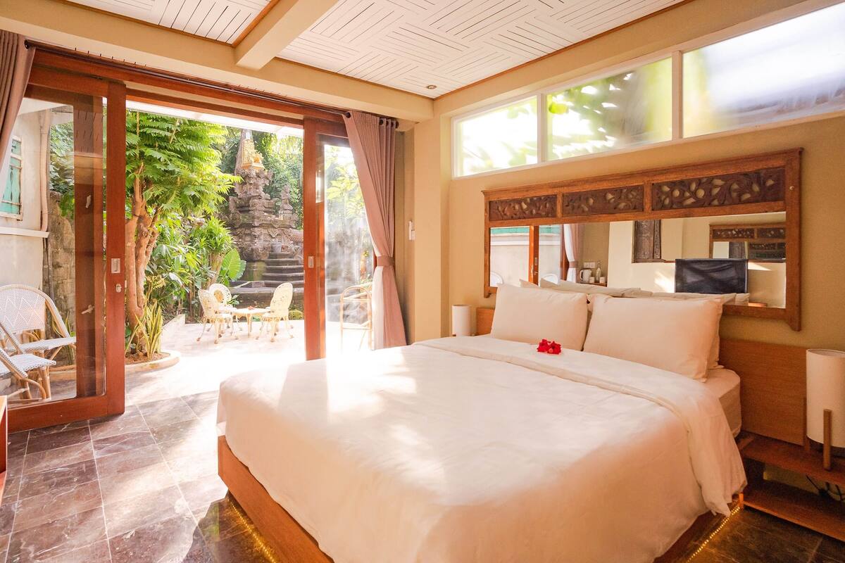 A spacious bedroom with a large bed, a wooden headboard, a television, and a view of the garden through an open door.