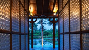 A corridor with a view of a palm tree and water, leading to a covered area with wooden beams and glass panels. - Nayaka Living Ubud (Ubud)