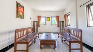 A room with two wooden benches, a central table, and windows with curtains. - Ganandra Stay Denpasar (Denpasar)