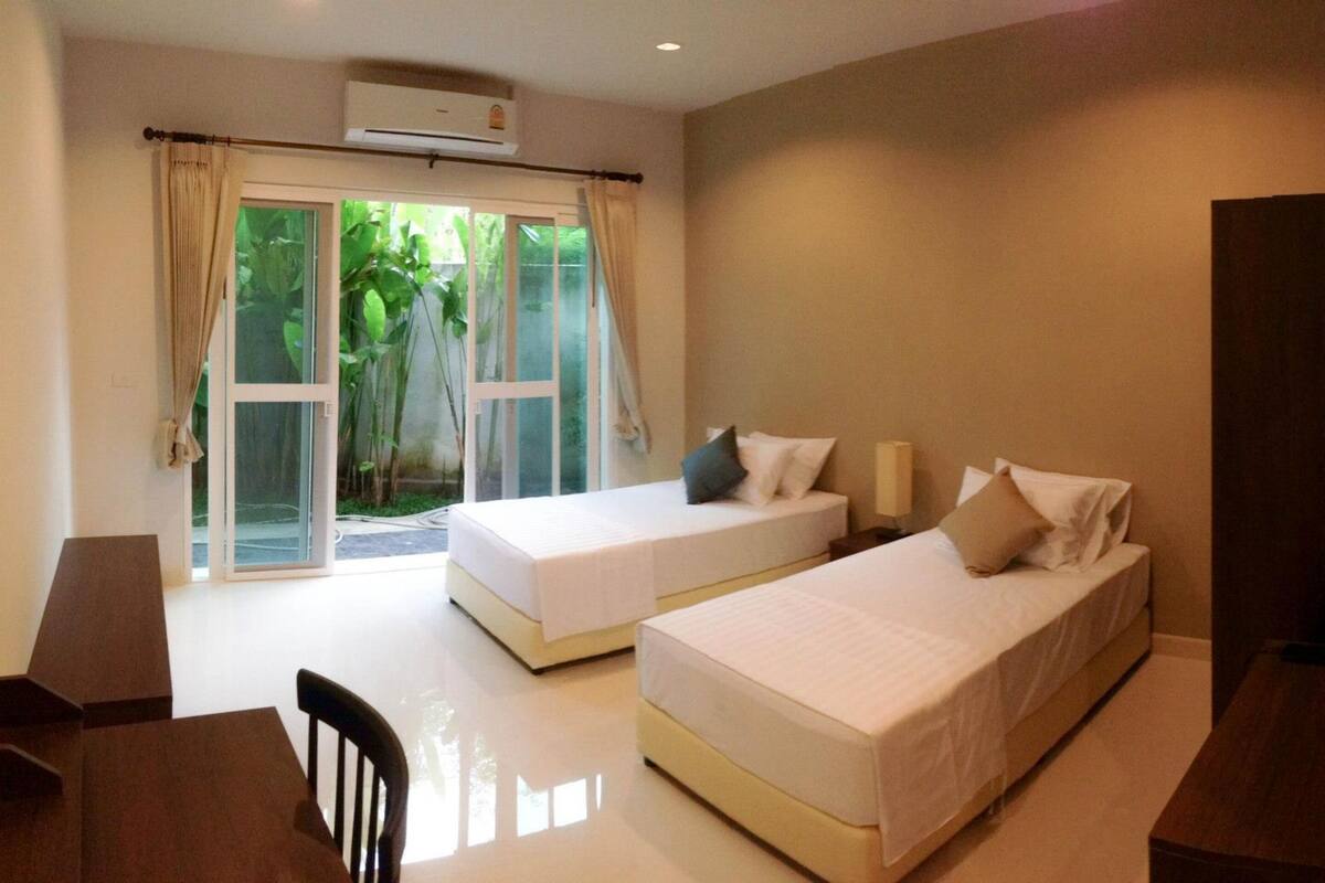 A hotel room with two beds, a desk, and a chair. There is a sliding glass door leading to a balcony with greenery.