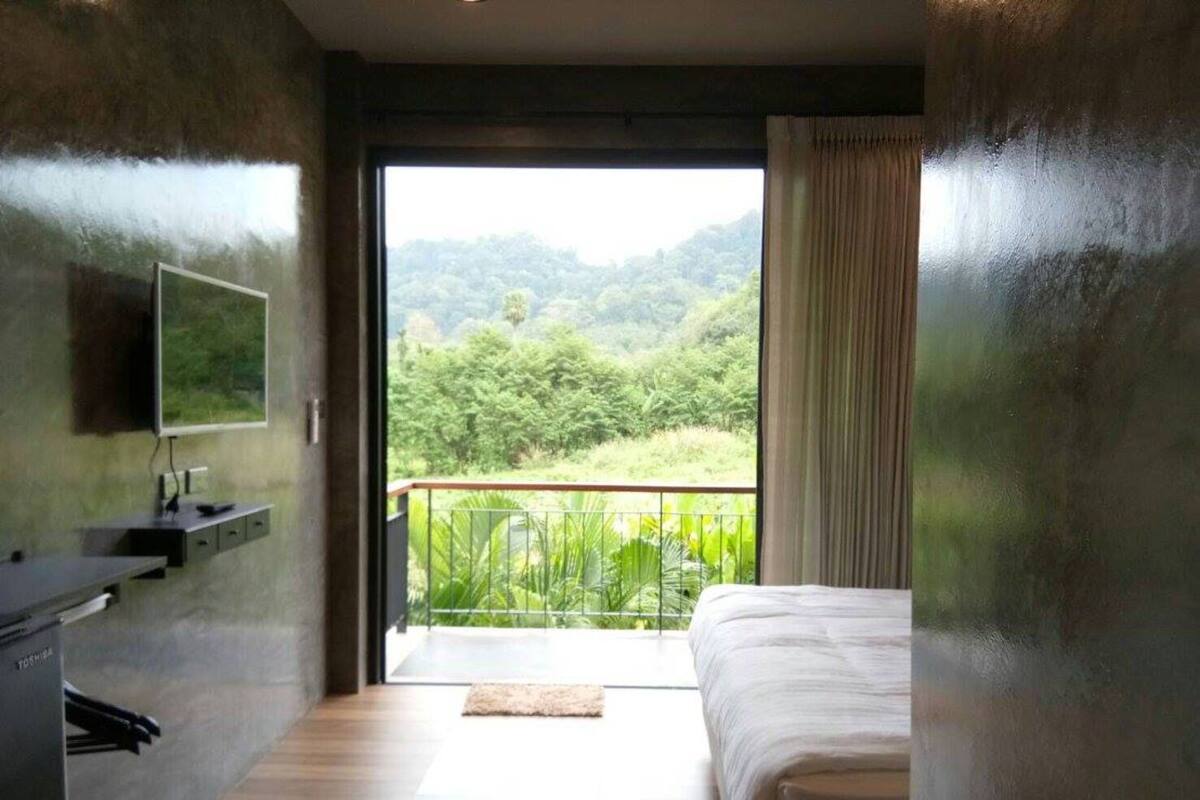 A bedroom with a bed, a desk, and a TV on the wall. There is a balcony with a view of greenery.