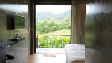 Quarto com cama, escrivaninha e televisão na parede. Há uma varanda com vista para o verde.
