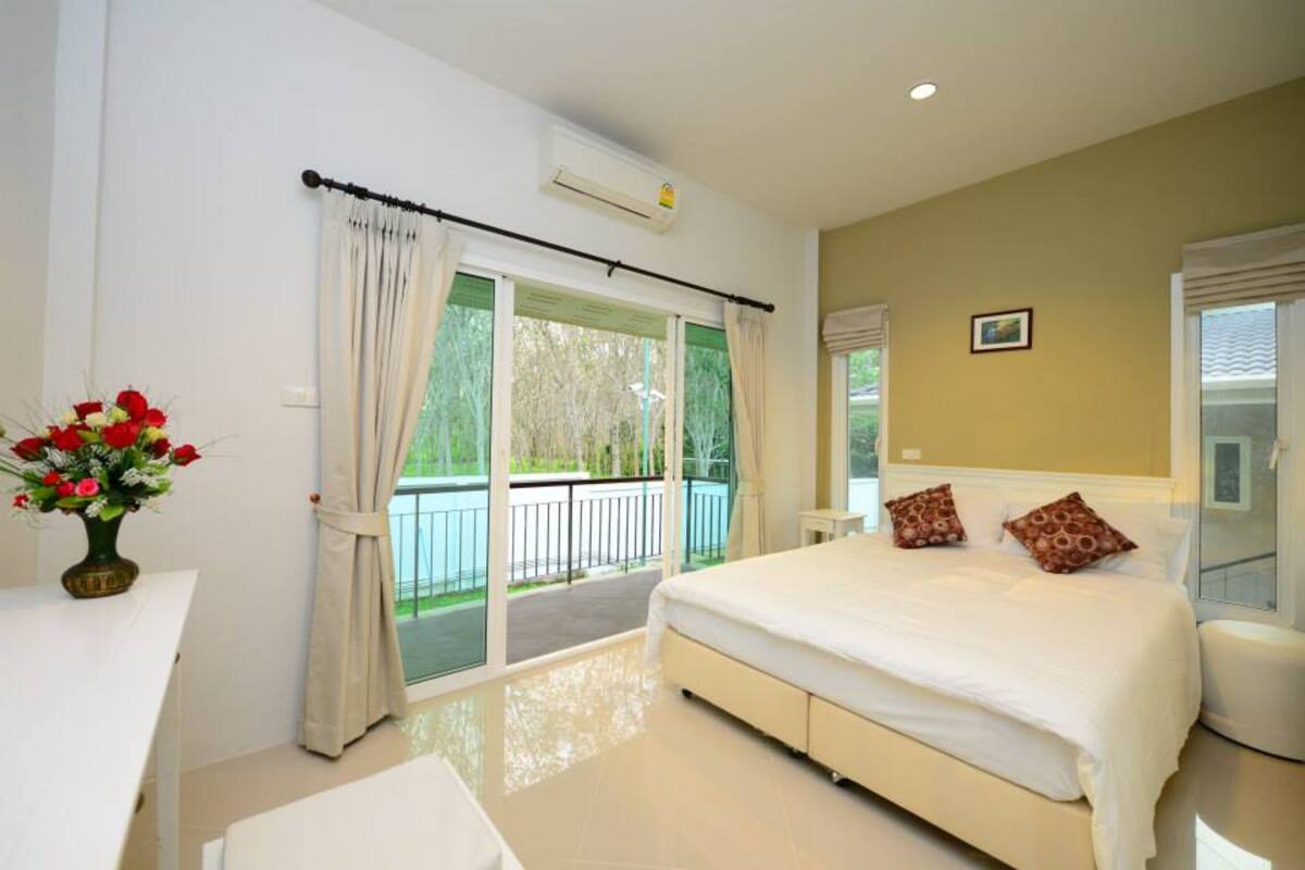 A bedroom with a large bed, a desk, and a chair. There is a window with curtains and a view of a balcony.