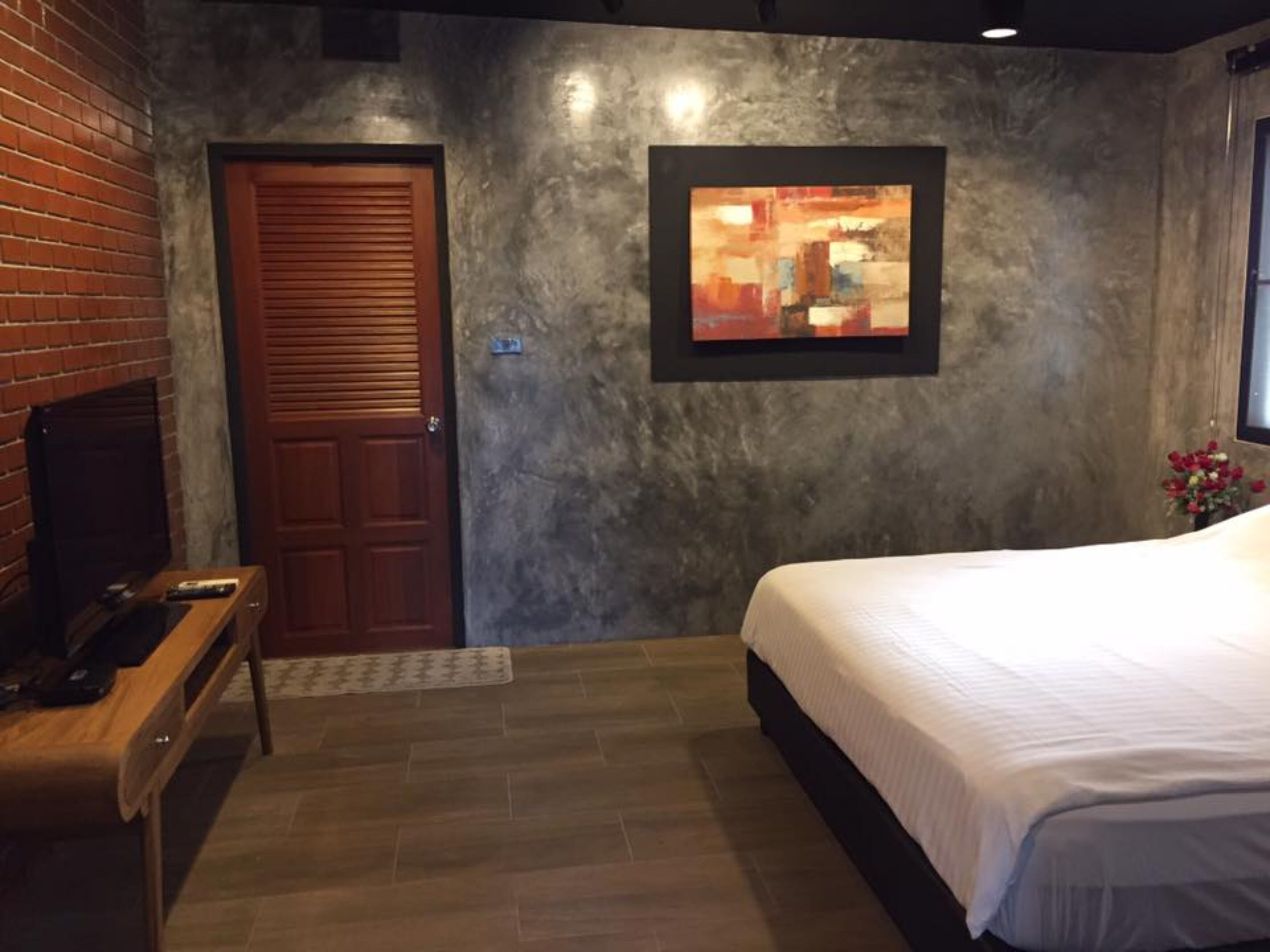 Un dormitorio con una puerta de madera, un cuadro abstracto enmarcado, una cama y una mesita con un control remoto.