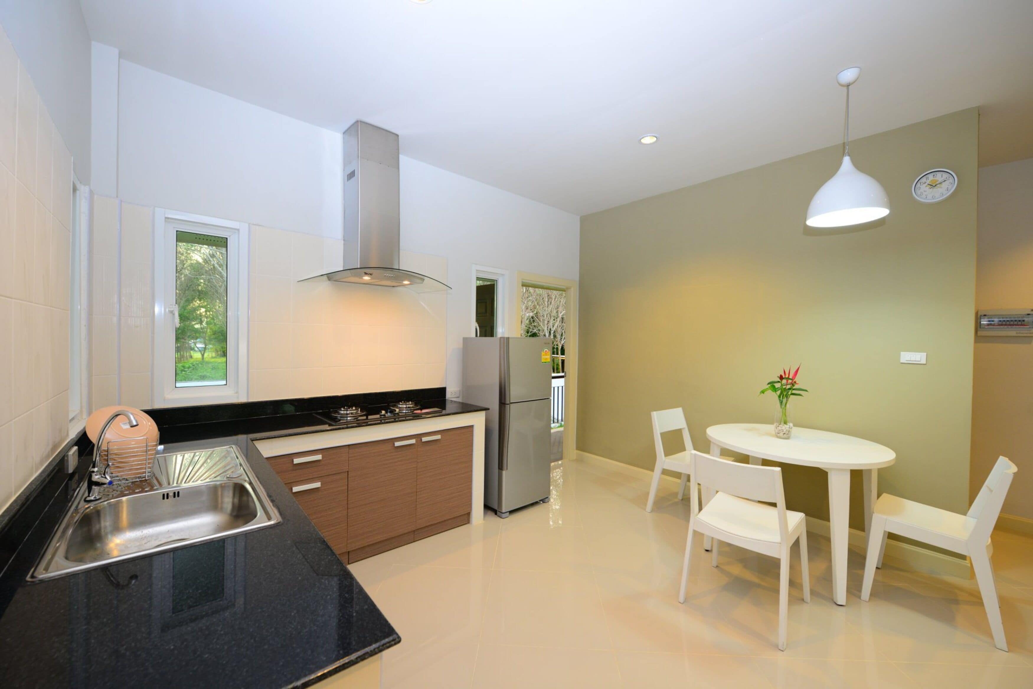 A modern kitchen with a stainless steel sink, a built-in oven, and a refrigerator. There is a dining area with a white table and chairs.