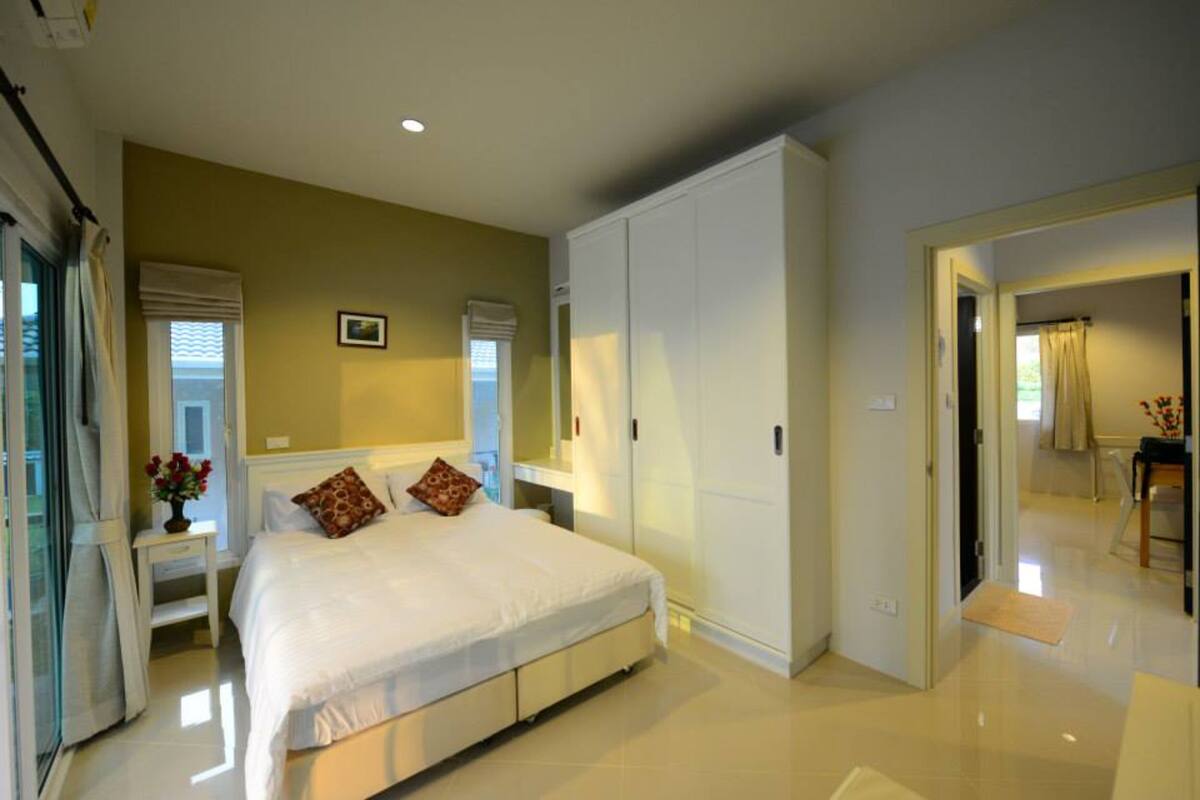 A bedroom with a large bed, a wardrobe, a bedside table with a vase of flowers, and a window with curtains.