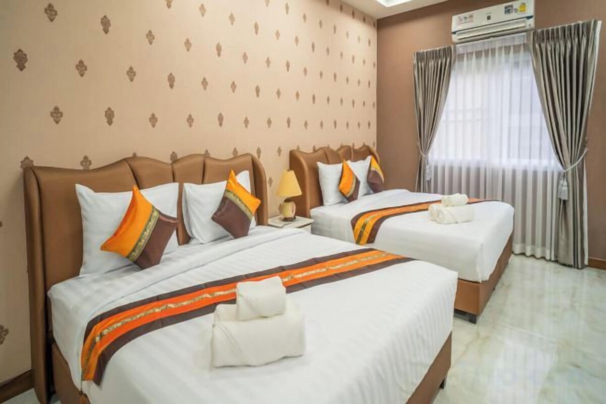 A hotel room with two beds, each with matching pillows and a blanket, a bedside table with a lamp, and a window with curtains.