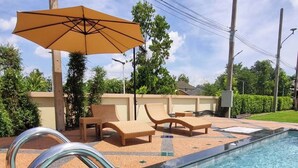 A swimming pool with lounge chairs and an umbrella, surrounded by a tiled deck and greenery.