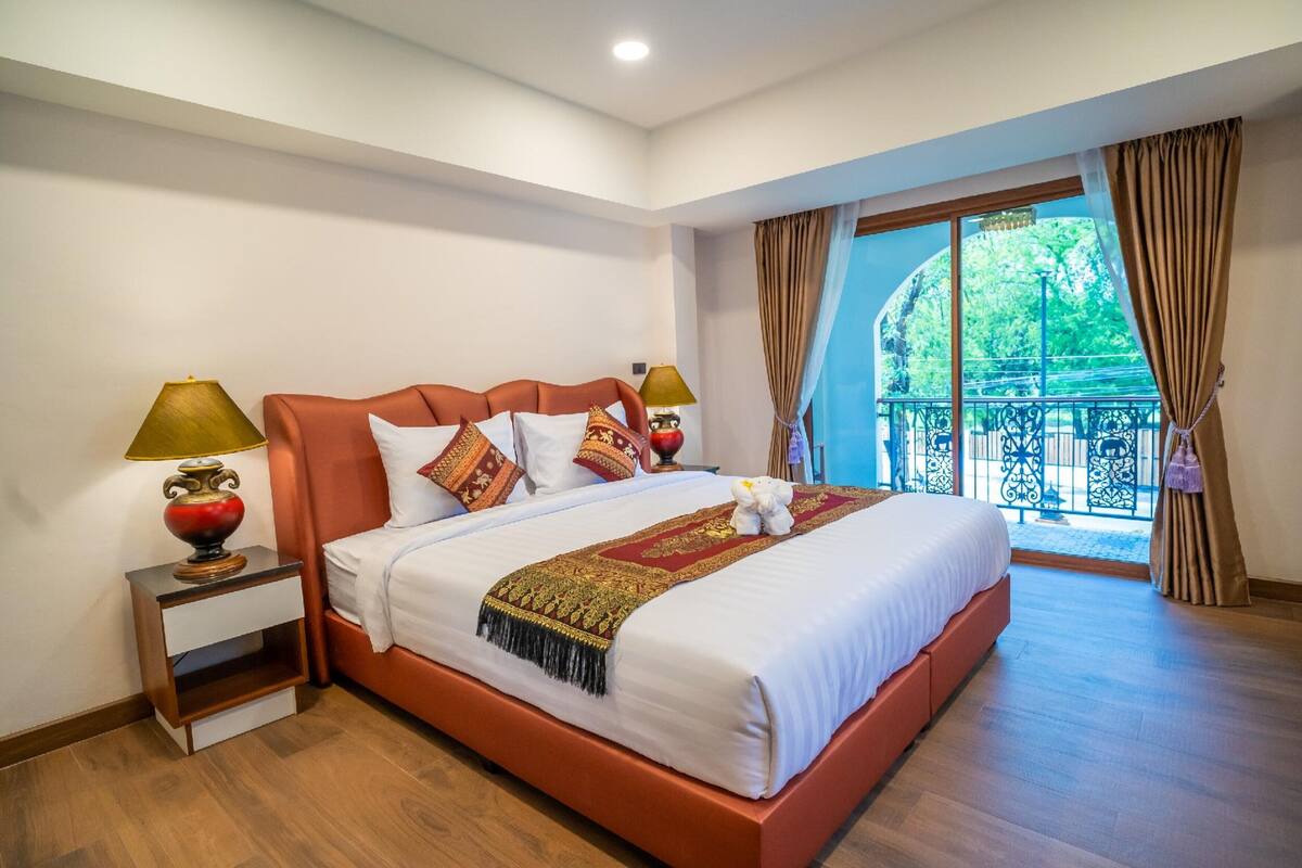 A spacious bedroom with a large bed, two bedside tables with lamps, a balcony with a view of greenery, and a window with curtains.