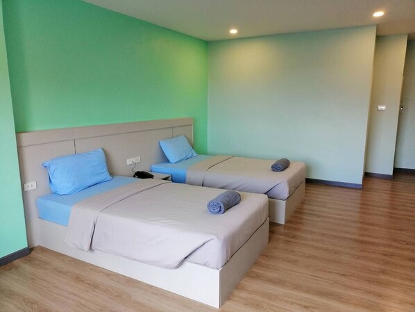 A hotel room with two beds, each with a headboard and pillows. There is a bedside table with a phone and a power outlet. The room has wooden flooring and light green walls.