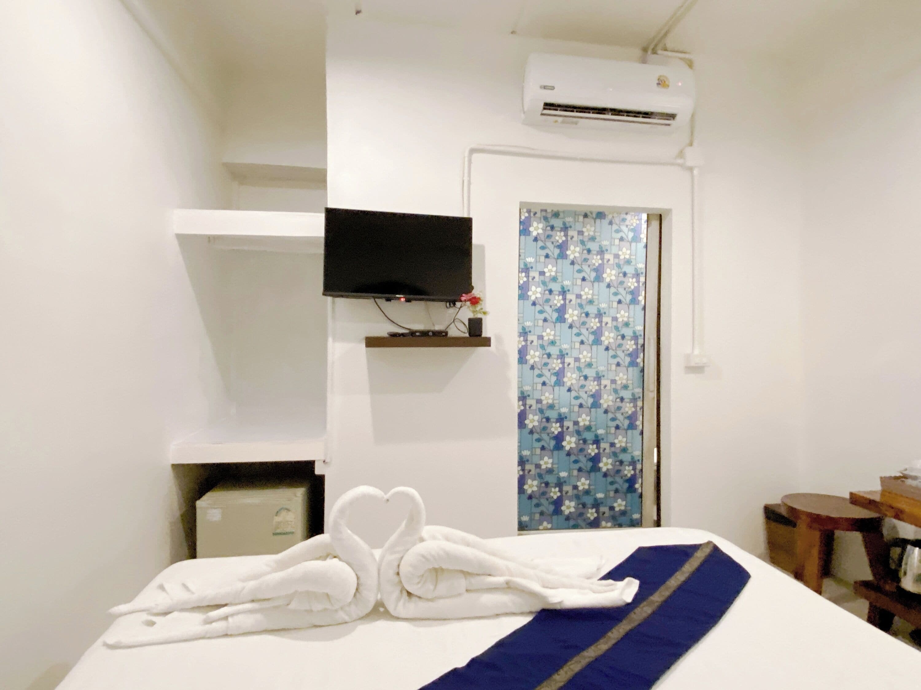 Une chambre d’hôtel comprenant un lit, un téléviseur à écran plat fixé au mur, un appareil de climatisation, une petite étagère ornée d’un objet décoratif et un rideau à motifs floraux.