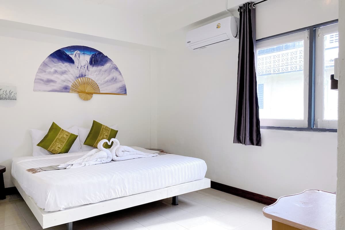 A neatly made bed with white linens, a wall-mounted air conditioner, a window with dark curtains, and a wall art of a horse and sun.