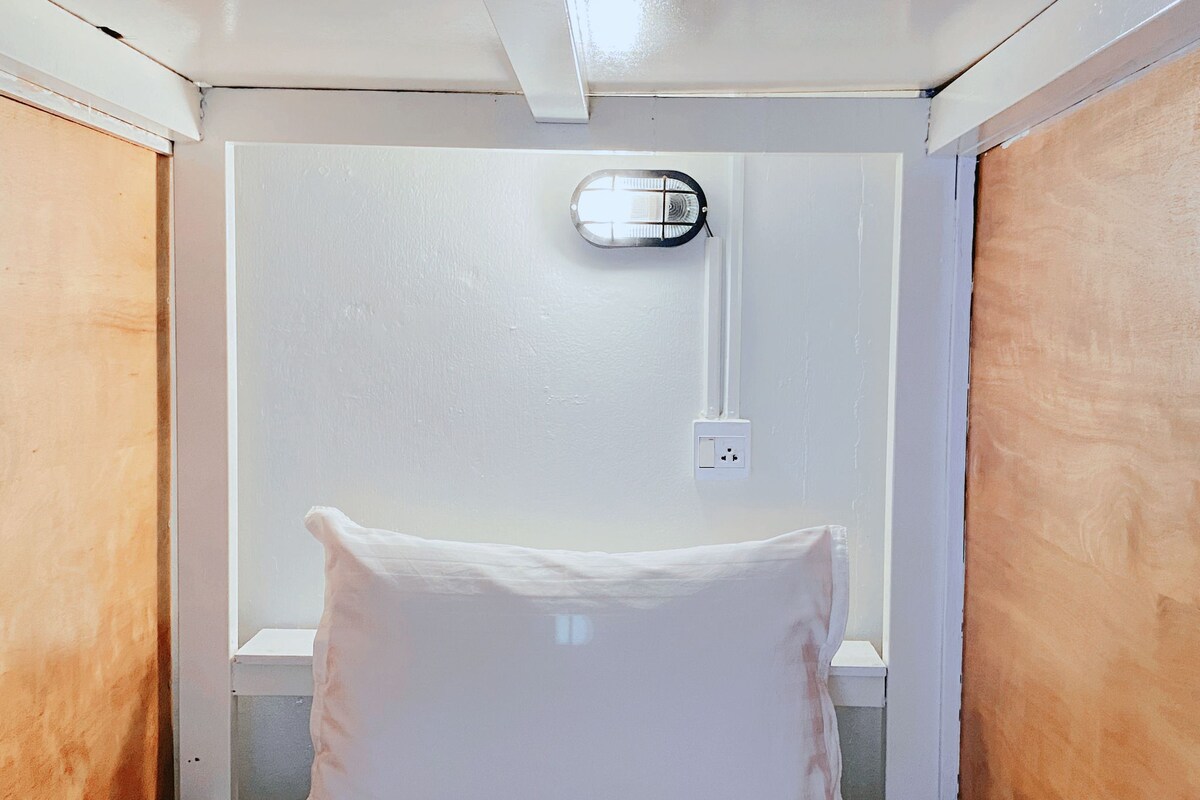 A small, single-bed room with a white wall, a ceiling light fixture, and a power outlet.