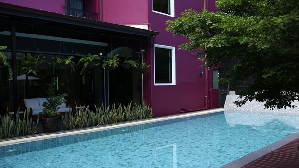 Una piscina con borde de azulejos, rodeada por una terraza de madera y un edificio morado con ventanas y una puerta.