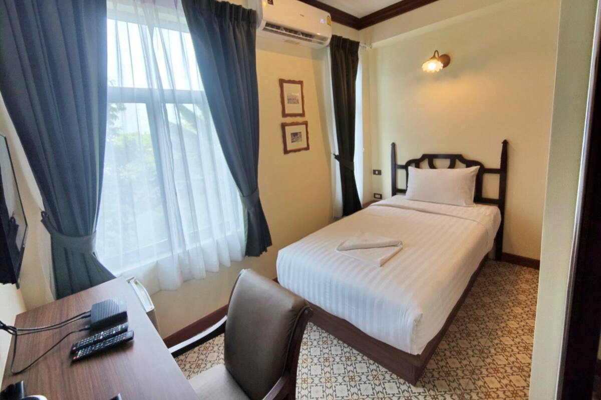 A hotel room with a bed, a desk with a chair, a window with curtains, an air conditioning unit, and a wall-mounted light fixture.