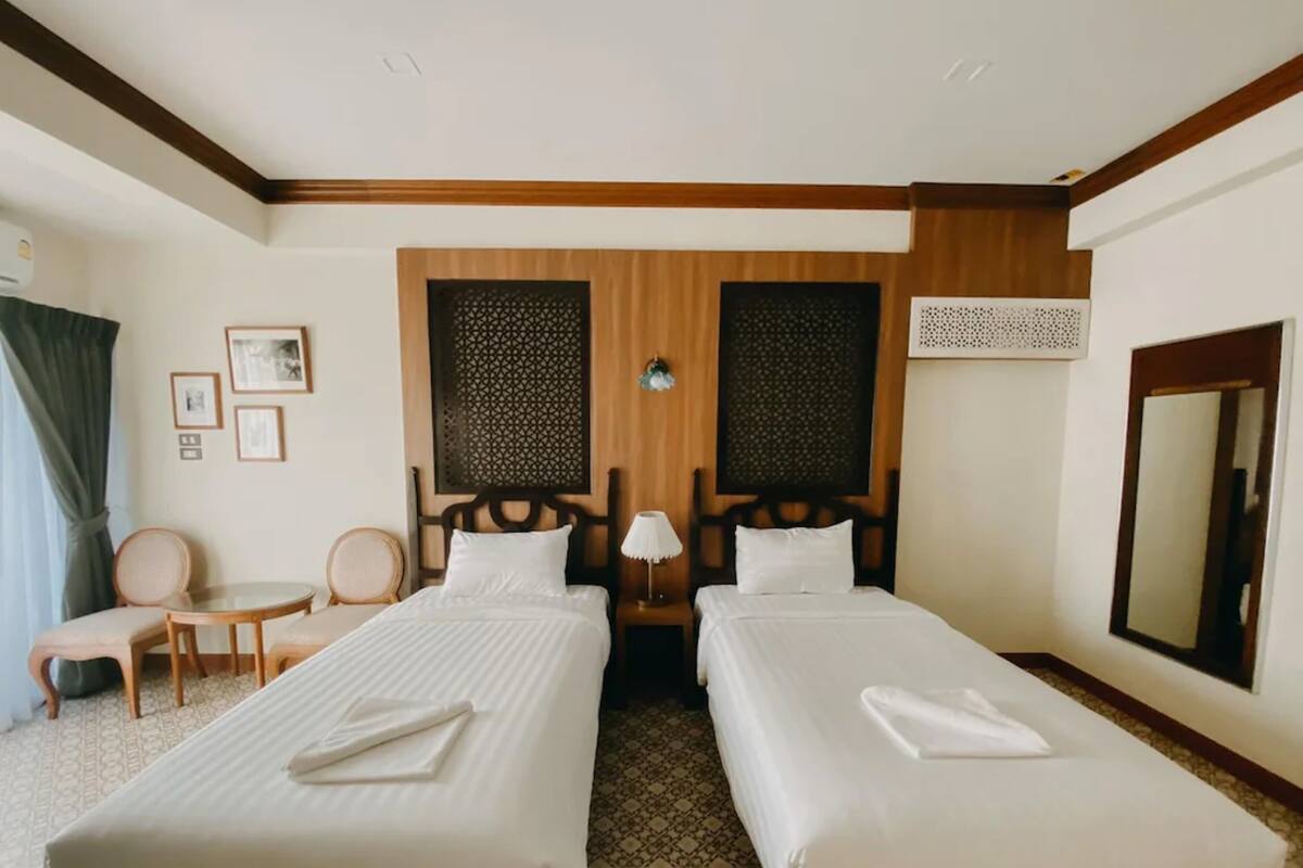 A hotel room with two single beds, a small round table, two chairs, a mirror, and decorative wall panels.