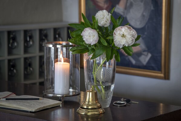 Una vela en un candelabro de vidrio, un florero con flores blancas, una campana de bronce, llaves y una libreta sobre una mesa de madera.