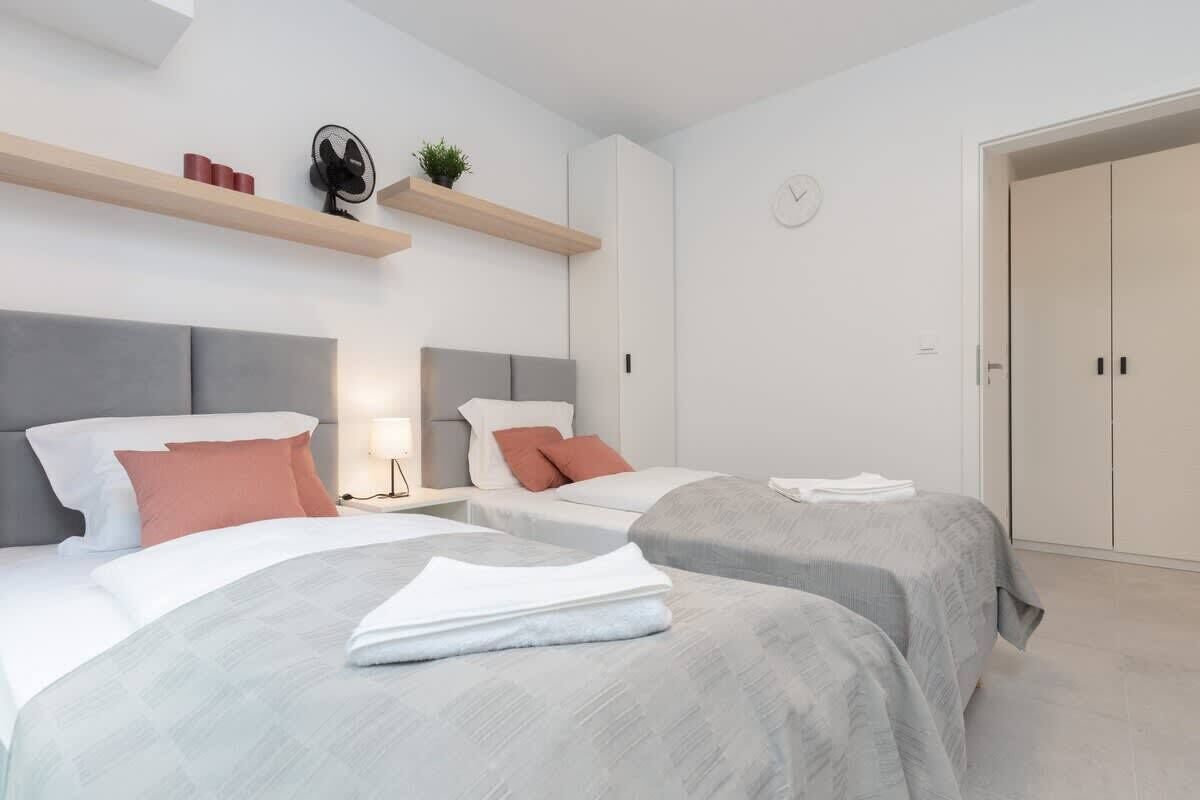 A modern hotel room with two beds, a bedside table with a lamp, a wall clock, and a wardrobe.