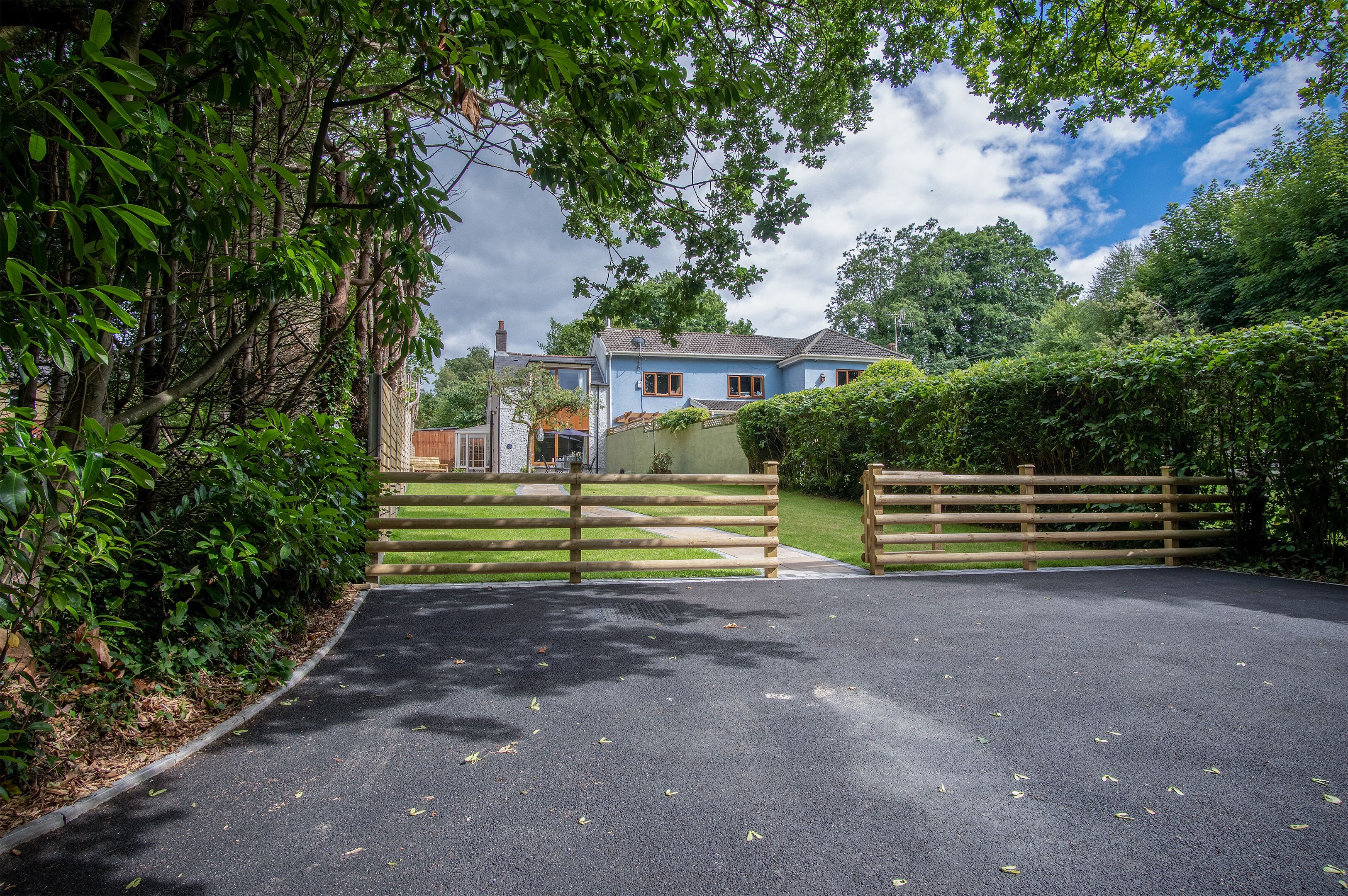A gated driveway leading to a house with a blue facade, surrounded by lush greenery and trees.