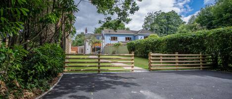 A gated driveway leading to a house with a blue facade, surrounded by lush greenery and trees.