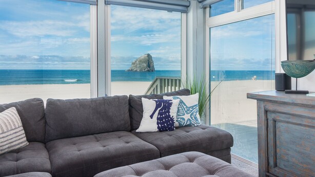 A modern living room with a gray sectional sofa, a small table, and a view of the ocean and a rock formation through large windows.