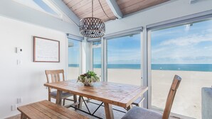 A wooden dining table with chairs and a bench, a hanging light fixture, and a view of the beach through large glass doors.