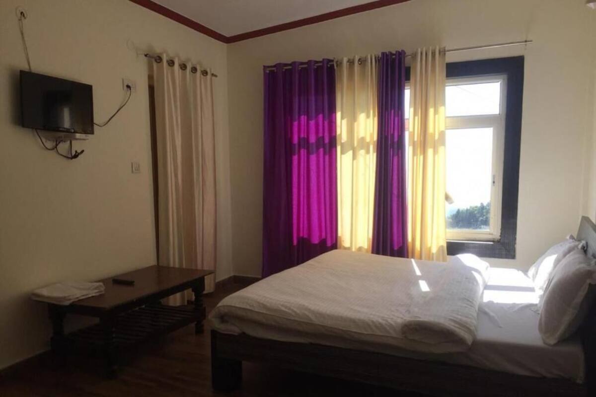 Un dormitorio con cama, una mesita, televisor montado en la pared y ventana con cortinas.