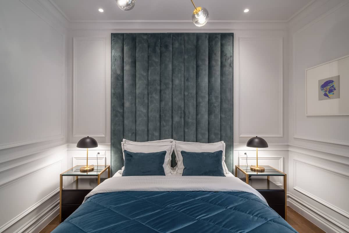 Un dormitorio moderno con una cama grande, dos mesitas de noche con lámparas, un cabecero gris y un cuadro enmarcado en la pared.