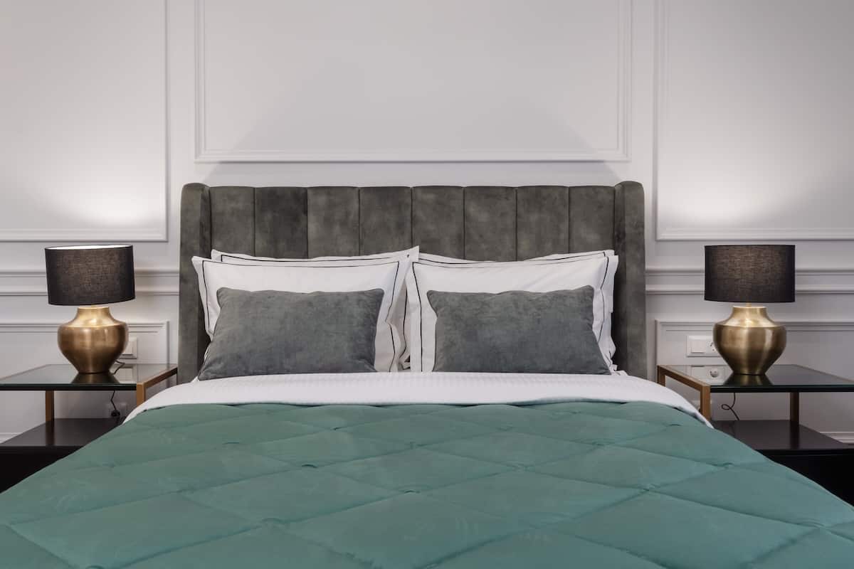 Una cama bien hecha con colcha verde azulada, dos almohadas y un cabecero tapizado gris. Hay dos mesitas de noche con lámparas a cada lado de la cama.