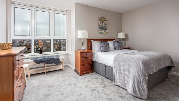 A bedroom with a bed, bedside tables, a dresser, a bench, and a window with a view of buildings. - Marlborough Court - Donnini Apartments (Ayr)