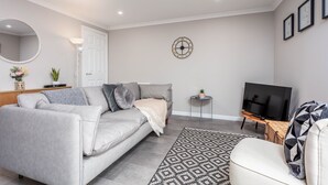 A modern living room with a grey sofa, a patterned rug, a television, and a wall clock. - Marlborough Court - Donnini Apartments (Ayr)