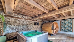 Un mur de pierres, un sauna en forme de tonneau en bois et un jacuzzi dans une pièce rustique aux poutres apparentes.
