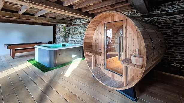 Un bain à remous en forme de tonneau, fait de bois, doté d’une porte en verre, d’un banc en bois et d’un mur en pierre, dans un décor rustique.