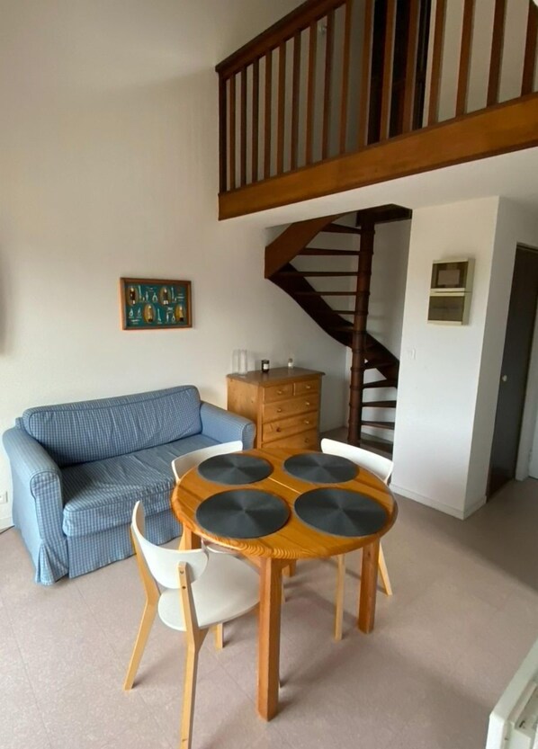 A living room with a blue sofa, a wooden dining table with four chairs, a staircase, and a wooden cabinet.