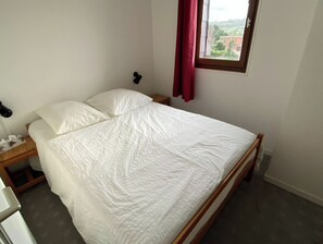 A bedroom with a bed, a nightstand, a lamp, a window with a curtain, and a view of buildings outside.