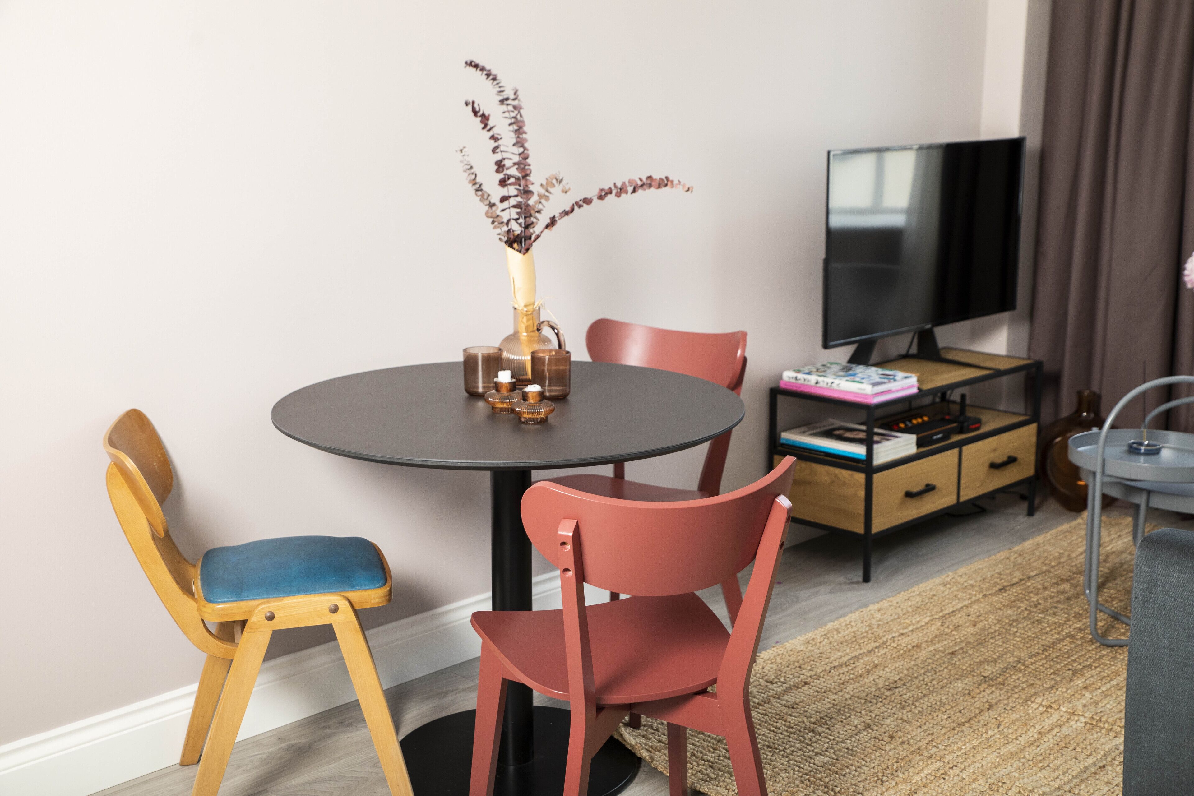 A dining area with a round table, two chairs, a vase with dried plants, a television, and a small cabinet with drawers.