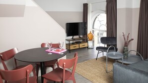Un salon moderne avec une table de salle à manger ronde, des chaises rouges, un téléviseur à écran plat, un canapé et une petite table d’appoint sur laquelle est posé un vase.