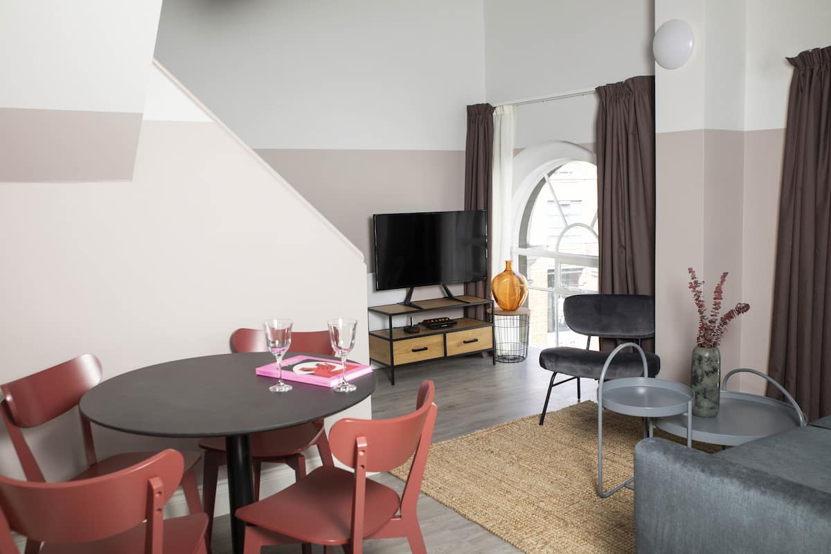Ett modernt vardagsrum med ett runt matbord, röda stolar, en platt-TV, en soffa och ett litet sidobord med en vas.
