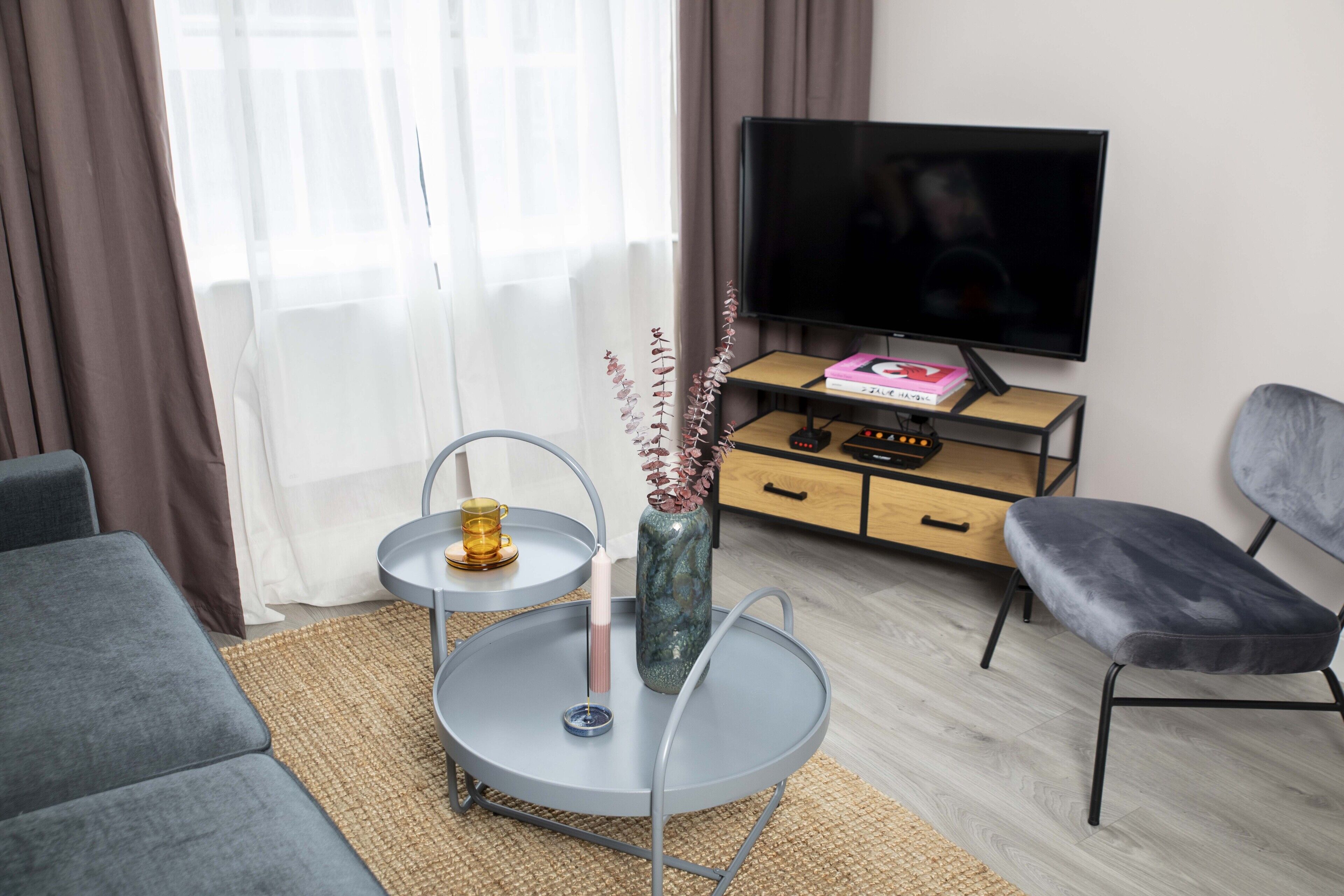 A modern living room with a grey sofa, a round coffee table with a vase and candle, a TV on a wooden stand, and a grey armchair.