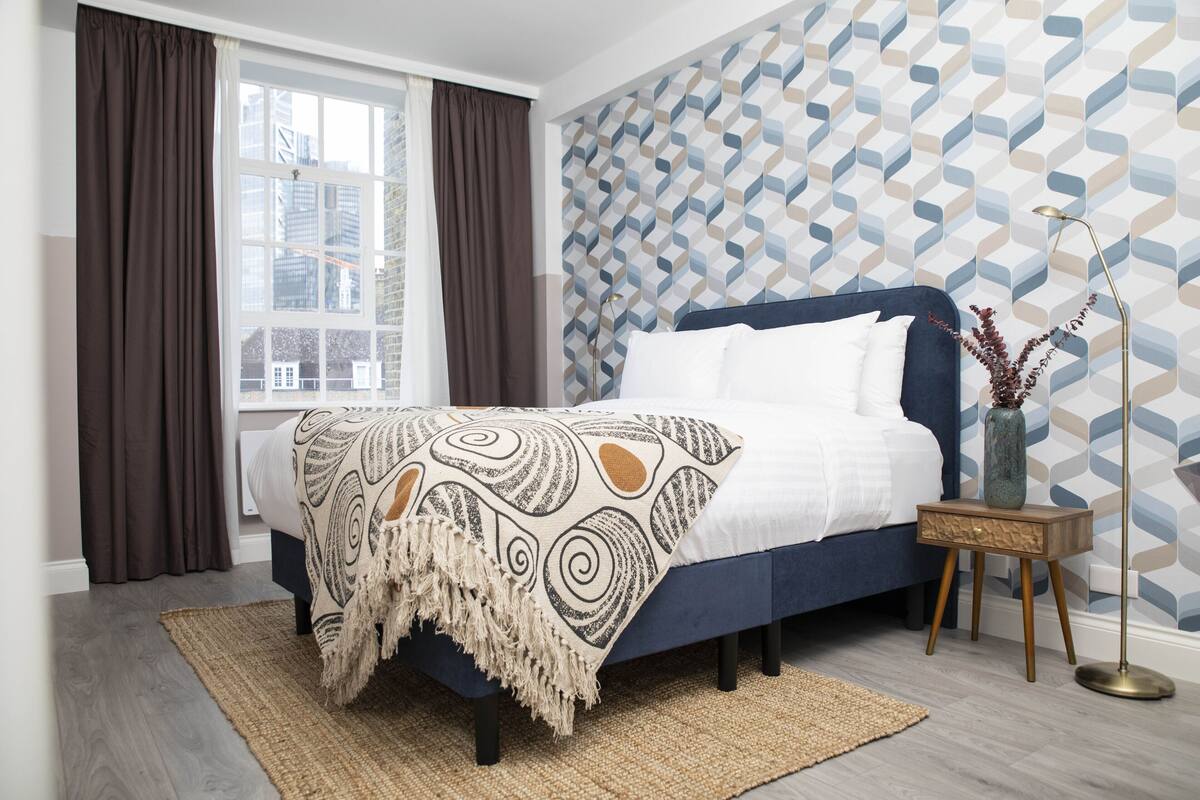 A bedroom with a bed, a window with curtains, a patterned wall, and a floor lamp.