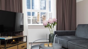 Un salon moderne avec un canapé gris, un téléviseur noir posé sur un meuble en bois, une petite table ronde ornée d’un vase de fleurs et une fenêtre avec des rideaux.