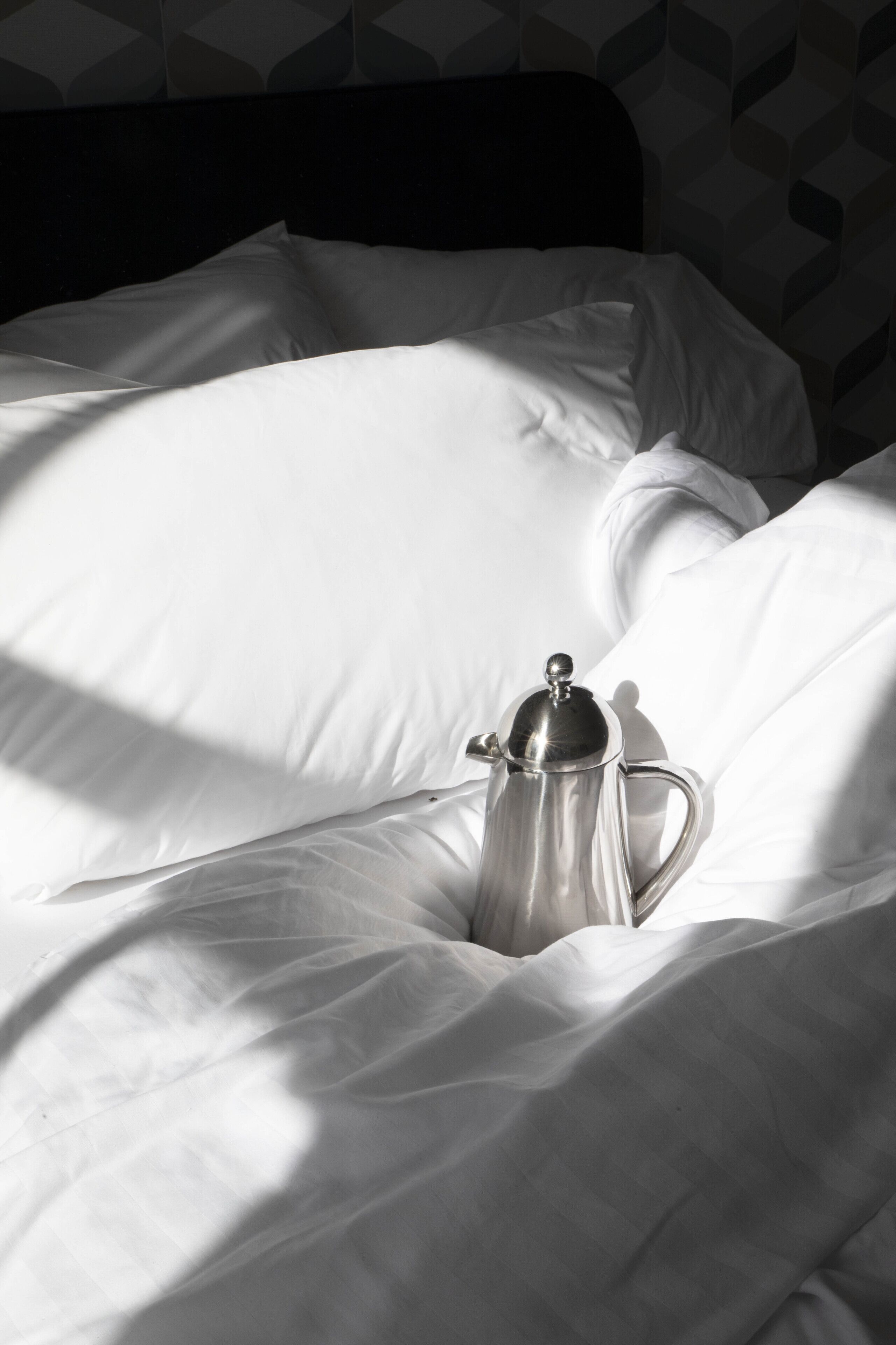 A bed with white linens and a silver coffee pot on it.