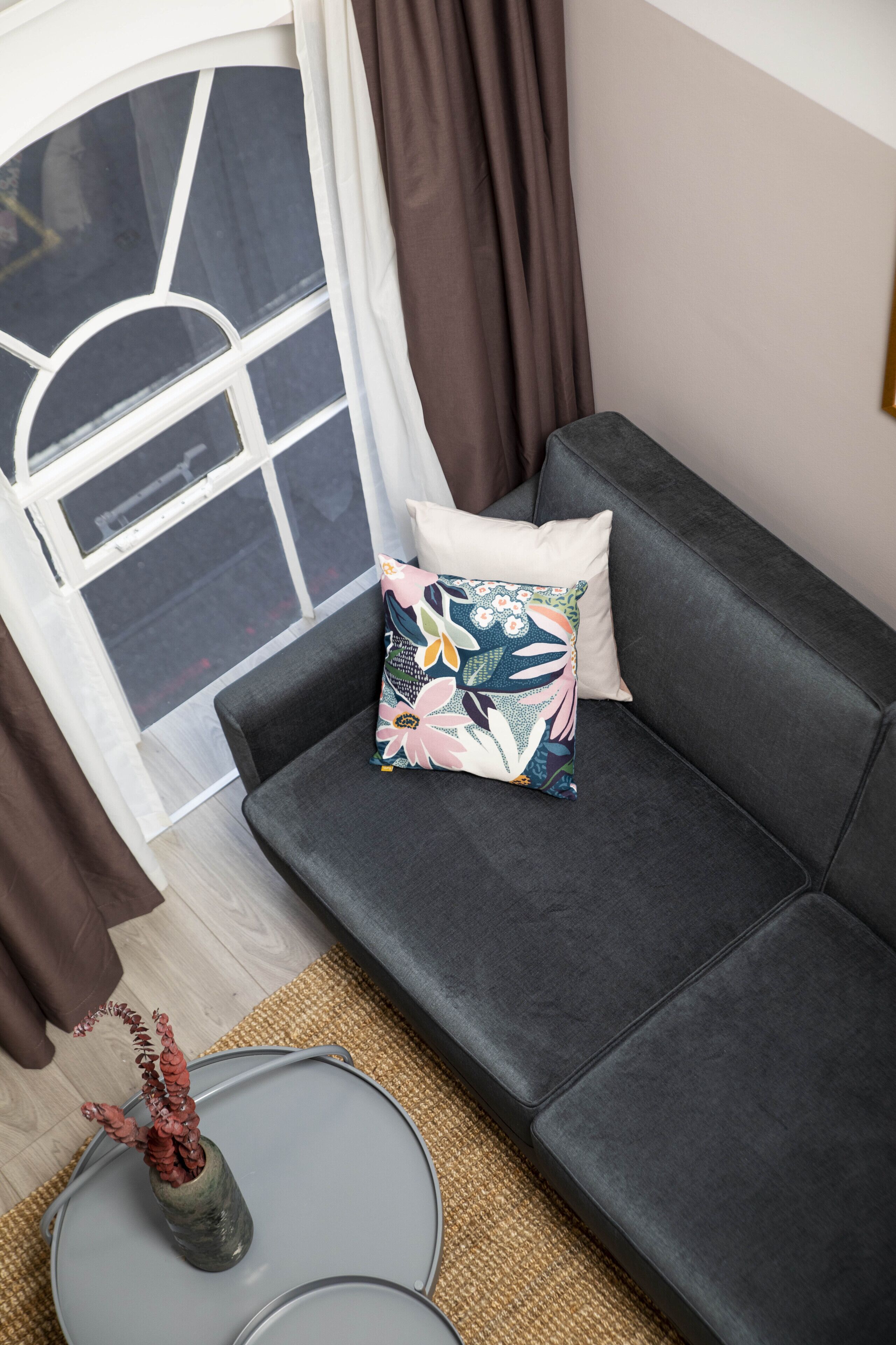 A modern living room with a dark grey sofa, a patterned pillow, a small grey table, and a vase with dried plants.