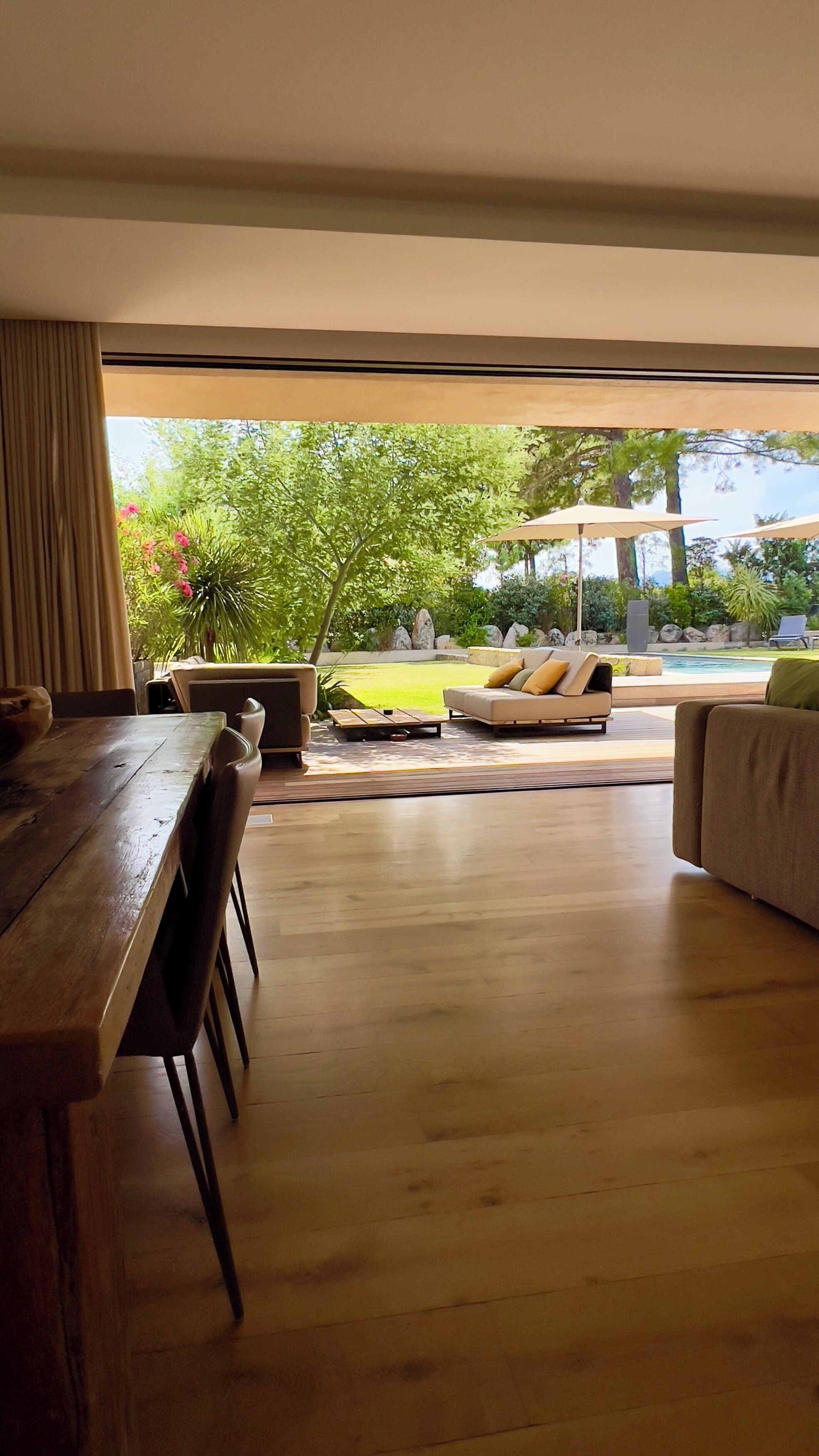 Een ruime woonkamer met een houten eettafel, een bank en een groot raam met uitzicht op een tuin en een terras.