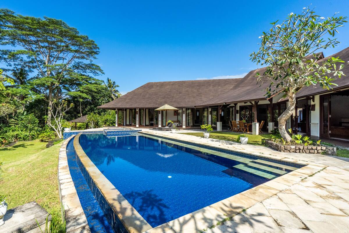 Piscina com deck de pedra, rodeada por um espaço coberto com área de estar e gramado.