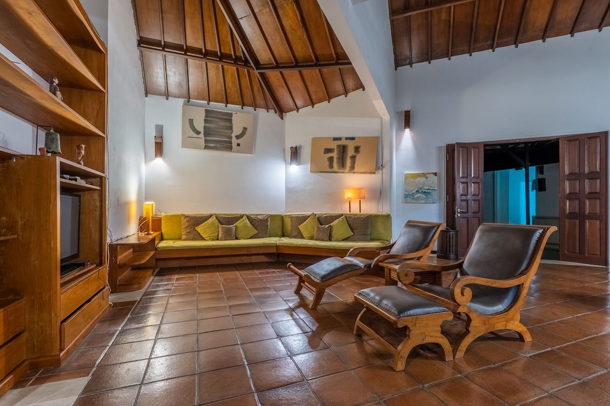 Uma sala de estar aconchegante com um sofá verde, cadeiras de madeira, piso ladrilhado e teto de madeira.